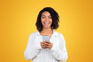 Chatting With Friends. Happy black woman holding mobile phone, looking at camera over yellow background, free space