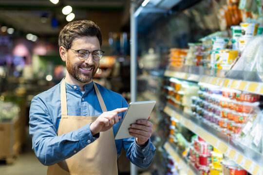 Confident grocery store worker uses digital tablet while standing in supermarket aisle. Surrounded by various products, he engages with technology to manage inventory. - Powered by Adobe