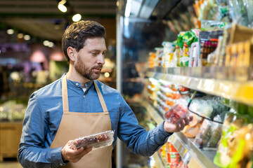 Man in grocery store aisle examining fresh produce while wearing blue shirt and apron. Holding packaged fruit, deciding on purchase, surrounded by variety of products under warm lights.
