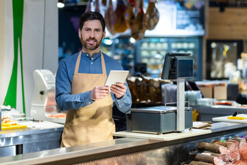 Professional shopkeeper in apron using tablet at delicatessen counter of modern grocery store. Warm...