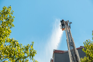 Rear view of firefighters extinguish fire on the roof of house using extended ladder equipped with water hose to spray water onto the building rooftop with smoke. Fireman extinguishing fire © samael334