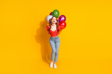 Full size photo of pretty young girl hold air balloons kissing lips wear trendy red outfit isolated on yellow color background