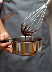 Fototapeta premium Man in grey apron with whisk and saucepan at kitchen, closeup photo of male hands. Man making chocolate. 