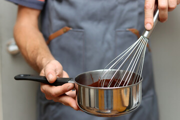 Man in grey apron with whisk and saucepan at kitchen, closeup photo of male hands. Man making chocolate. 