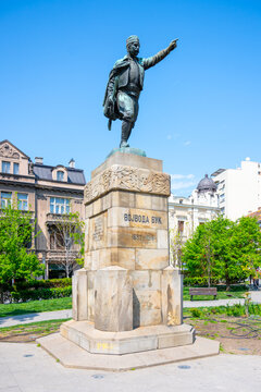 The Vojvoda Vuk statue stands prominently in Belgrade, Serbia, showcasing its detailed design against a backdrop of trees and modern architecture under a bright blue sky.