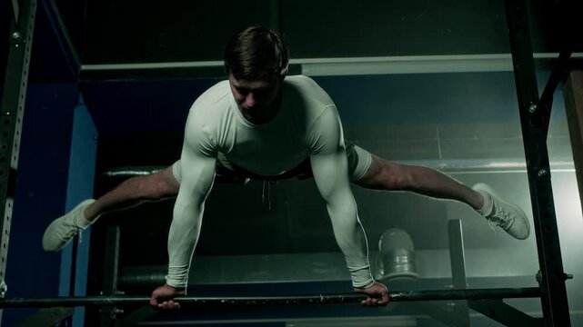 Flexible Caucasian young man gripping gym bar and doing straddle planche exercise during gymnastics training indoors