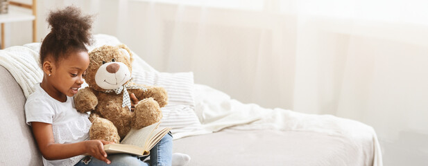 A young black girl sits comfortably on a sofa, engrossed in a book. Next to her, a plush teddy bear...