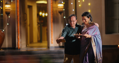 Indian Asian Hindu happy elder old gen x couple hold phone use cam look cute take pose selfie shoot video decor light home older aged male man female wife lady mom dad enjoy fun joy Diwali night time