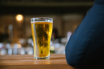 A man pours frothy beer into a glass at a lively pub. Friends gather around the bar, enjoying drinks and celebrating, with a warm, golden glow highlighting the joyful, friendly atmosphere.