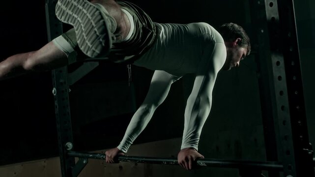 Lowkey shot of Caucasian young man in white long sleeved compression shirt performing straddle planche on uneven bar in dark gym