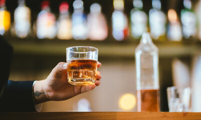 A stylish man holds a glass of whiskey in a fancy pub amidst a crowd of celebrants. The warm and elegant atmosphere accentuates the rich amber-colored liquid.