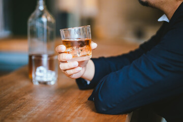 A stylish man holds a glass of whiskey in a fancy pub amidst a crowd of celebrants. The warm and elegant atmosphere accentuates the rich amber-colored liquid.