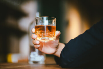 A stylish man holds a glass of whiskey in a fancy pub amidst a crowd of celebrants. The warm and elegant atmosphere accentuates the rich amber-colored liquid.