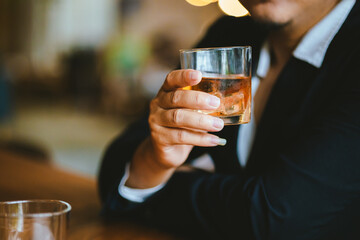 A stylish man holds a glass of whiskey in a fancy pub amidst a crowd of celebrants. The warm and elegant atmosphere accentuates the rich amber-colored liquid.