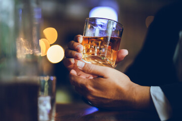 A stylish man holds a glass of whiskey in a fancy pub amidst a crowd of celebrants. The warm and elegant atmosphere accentuates the rich amber-colored liquid.