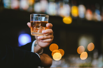 A stylish man holds a glass of whiskey in a fancy pub amidst a crowd of celebrants. The warm and elegant atmosphere accentuates the rich amber-colored liquid.