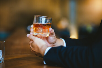 A stylish man holds a glass of whiskey in a fancy pub amidst a crowd of celebrants. The warm and elegant atmosphere accentuates the rich amber-colored liquid.