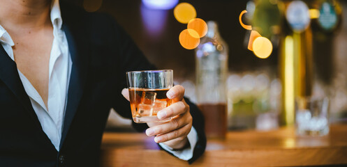 A stylish man holds a glass of whiskey in a fancy pub amidst a crowd of celebrants. The warm and elegant atmosphere accentuates the rich amber-colored liquid.