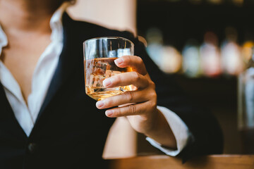 A stylish man holds a glass of whiskey in a fancy pub amidst a crowd of celebrants. The warm and elegant atmosphere accentuates the rich amber-colored liquid.