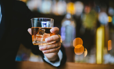 A stylish man holds a glass of whiskey in a fancy pub amidst a crowd of celebrants. The warm and elegant atmosphere accentuates the rich amber-colored liquid.