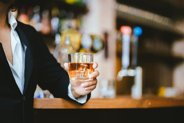 A stylish man holds a glass of whiskey in a fancy pub amidst a crowd of celebrants. The warm and elegant atmosphere accentuates the rich amber-colored liquid.