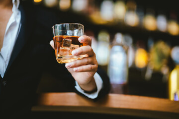 A stylish man holds a glass of whiskey in a fancy pub amidst a crowd of celebrants. The warm and elegant atmosphere accentuates the rich amber-colored liquid.