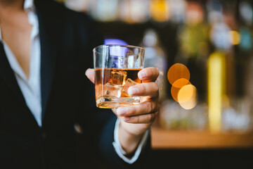 A stylish man holds a glass of whiskey in a fancy pub amidst a crowd of celebrants. The warm and elegant atmosphere accentuates the rich amber-colored liquid.