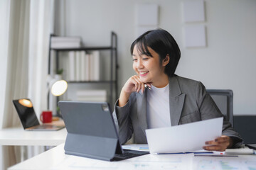 Young businesswoman is smiling while working from her modern office using a tablet and reviewing paperwork