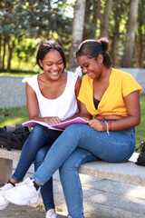 Two young Hispanic women sitting on a park bench, studying together from a shared notebook, smiling and enjoying their time learning in a natural setting, vertical image