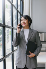 Businesswoman is holding a laptop and talking on the phone while standing near a window in an office building