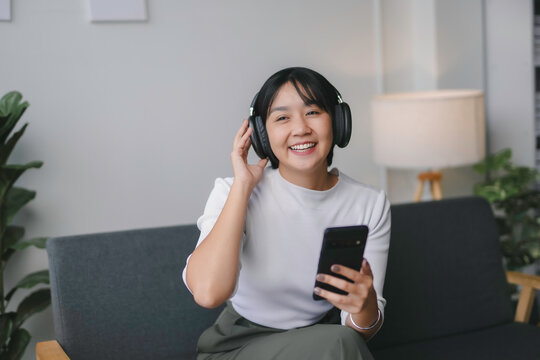 Happy young asian woman relaxing on sofa at home, enjoying listening to music using wireless headphones and holding smartphone in living room