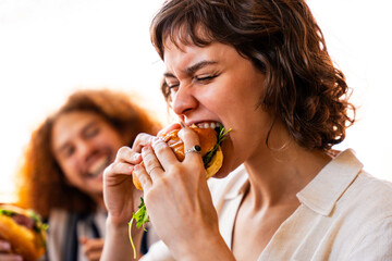 Multiethnic group of friends having meal at 80s vintage diner restaurant, woman biting cheeseburger...