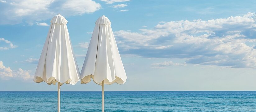 Close up of two closed white canvas beach umbrellas set against a backdrop of the sea and a blue sky with clouds. with copy space image. Place for adding text or design