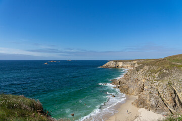 Fototapeta premium La côte sauvage du Finistère nord en Bretagne offre une plage de sable bordée de falaises escarpées, baignée par les eaux turquoises de la mer d'Iroise, sous un ciel bleu éclatant.