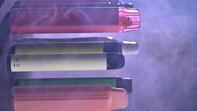A set of colorful disposable electronic cigarettes of different shapes on a black background with smoke. Concept of modern smoking, vaping and nicotine.