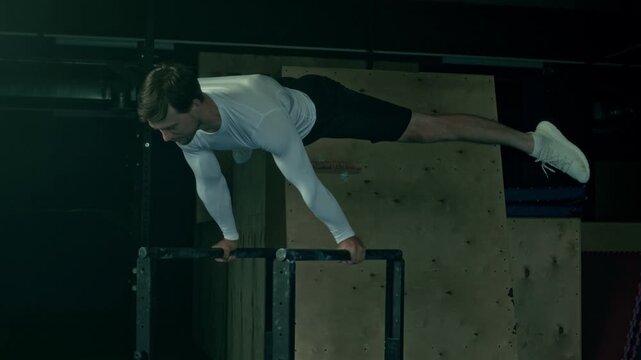 Athletic young man doing perfect straddle planche on bars while practicing indoors at dark loft style studio