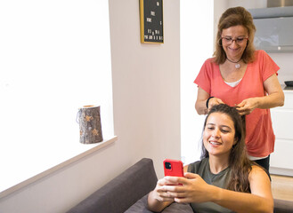 Smiling attractive older mother giving a nice braid hairstyle to her young beautiful brunette daughter who is sitting on the sofa with her cell phone in her hands.