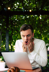 Surprised man reacting to something shocking while working on a laptop in a café with green plants around