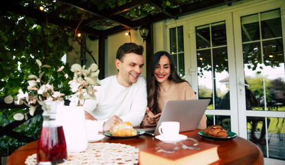 Coworkers work together with laptop outdoors. Two smiling individuals are engaged in conversation over drinks and pastries at a sunlit café. Freelancers discuss new project