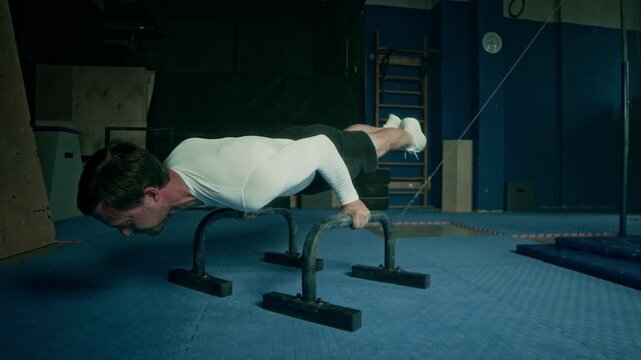 Acrobatic young Caucasian man with strong fit body under white long-sleeved compression shirt doing handstand and straddle planche exercises on two parallel bars in dark gym
