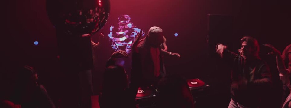 A crowd of people dancing on the dance floor of a nightclub under the light of colored spotlights. DJ mixing up the beats
