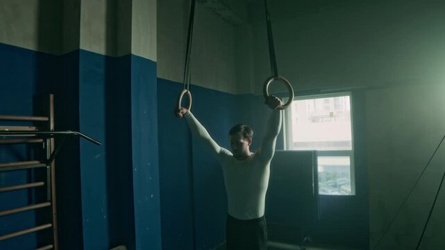 Athletic man practicing L sit and straddle planche exercises while training on gymnastics rings in dark gym
