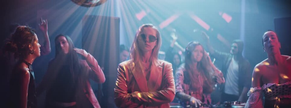 Close-up portrait of young woman looking at the camera and standing in the middle of a party surrounded by crowd, they are dancing around her