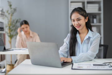 Smiling asian businesswoman working on laptop in modern office with colleagues working in background