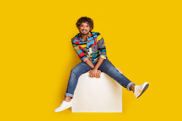 Full length photo of handsome young male sit white cube dressed stylish colorful garment isolated on yellow color background