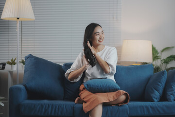 Obraz premium Young woman is sitting on a sofa in her living room, touching her hair and smiling