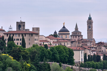 A panoramic view of upper town Bergamo’s historic center at dusk, featuring ancient towers, domes, and rooftops, with the misty hills in the background, capturing the charm of this Italian city.