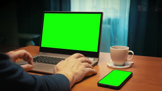A person using laptop working with Green Screen Display. User's hand scrolling touchpad.On the right is a phone with Green Screen Display and a cup of coffee. Close-up. Template for Content Creation.