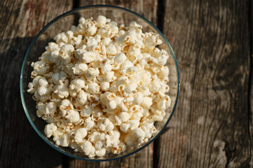 A glass bowl with fresh crispy fragrant salty popcorn stands on a wooden table on a sunny summer day. Top close up view. A classic snack for watching a movie or TV series.