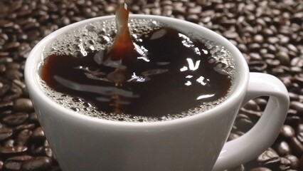 Close up of coffee bean drop in to hot espresso with pile of coffee seed. Macro of freshly roasted bean falling in to cup with aromatic smoke. Slow motion. Organic coffee seed concept. Comestible.